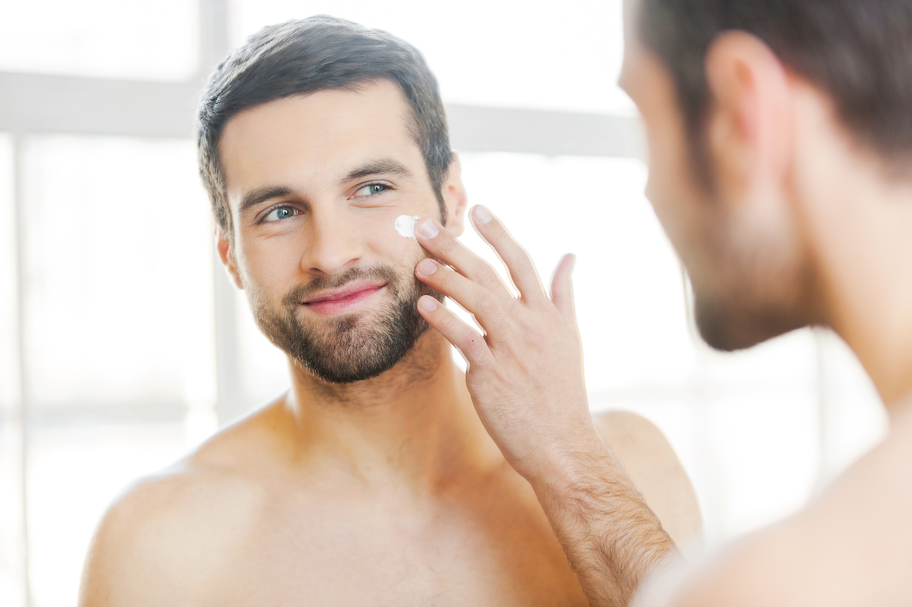 man applying skincare product to face in mirror